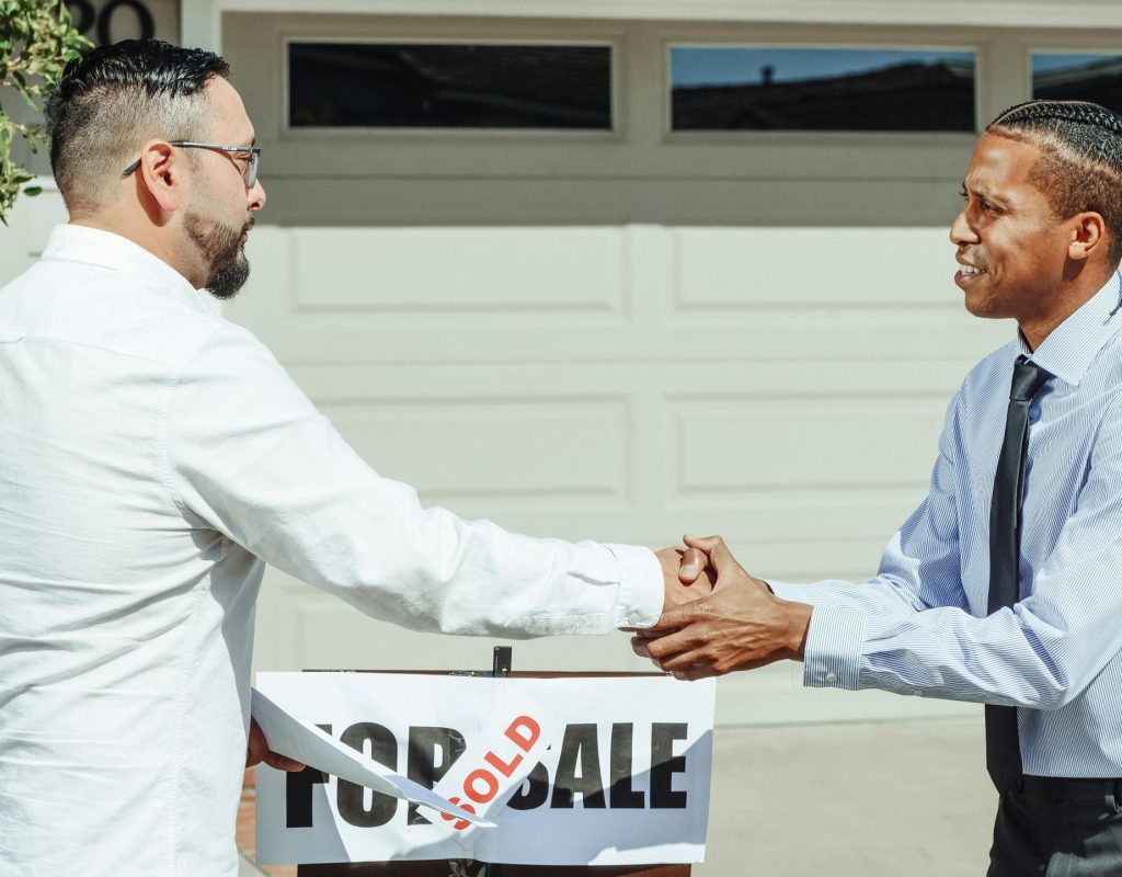 Two men shaking hands in front of a sold house sign, sealing a real estate transaction.