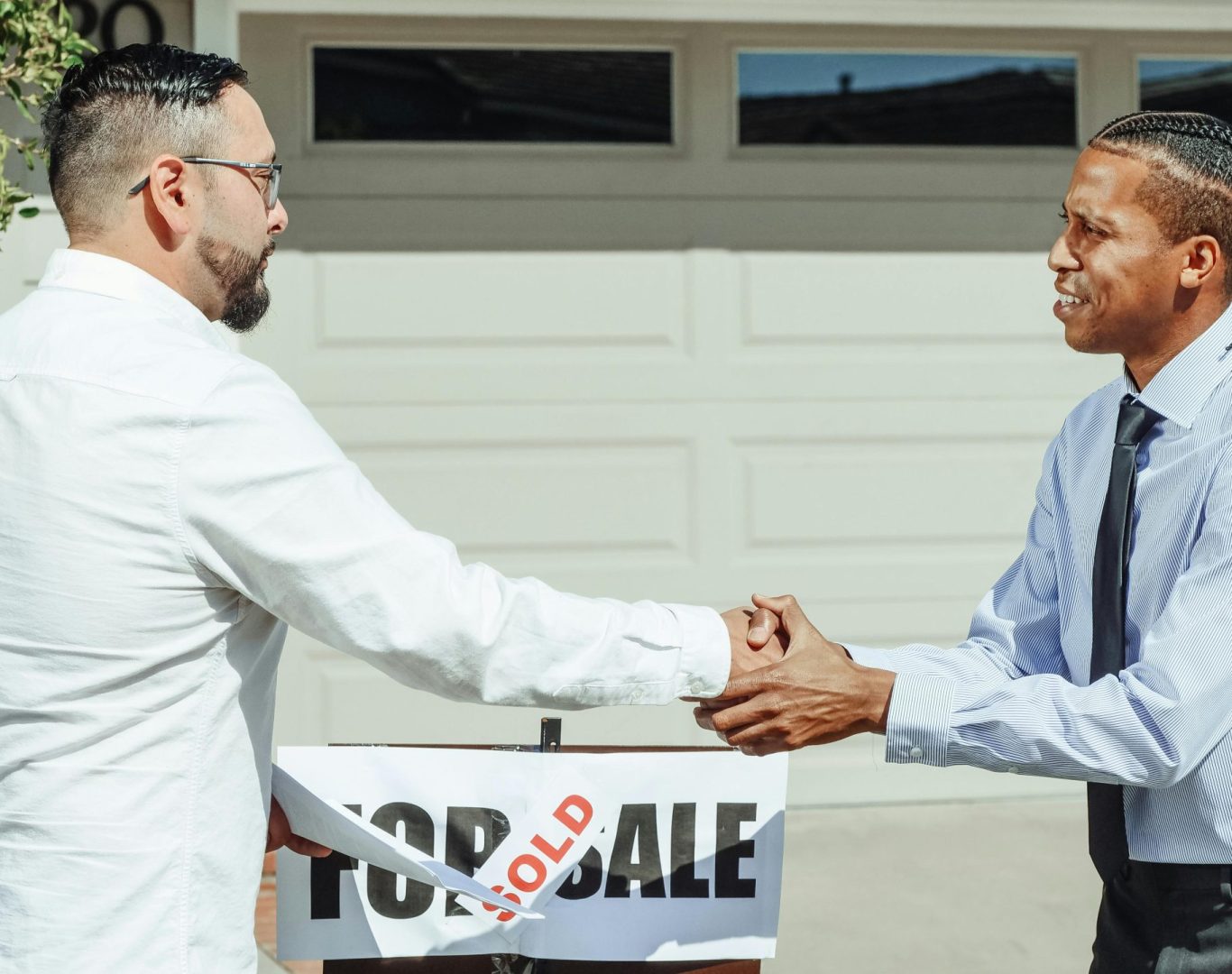 Two men shaking hands in front of a sold house sign, sealing a real estate transaction.