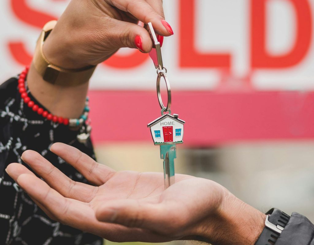Close-up of hands exchanging house key with colorful SOLD sign in the background.
