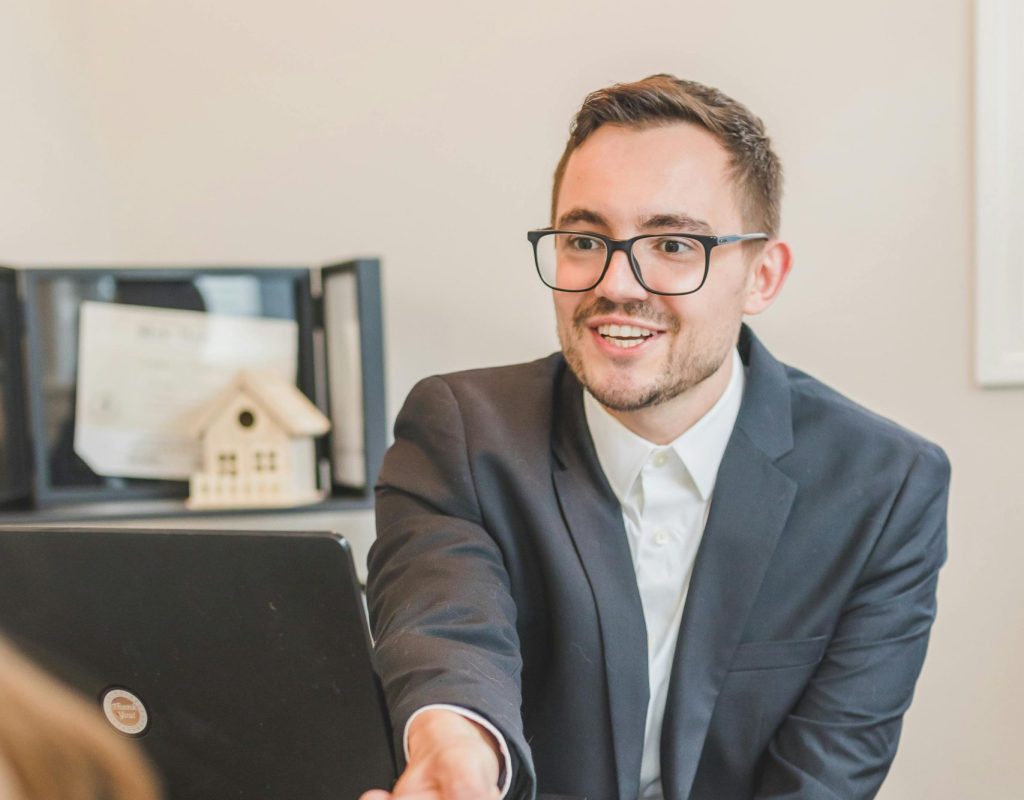 A professional real estate agent welcomes a client in the office with documents on the desk.