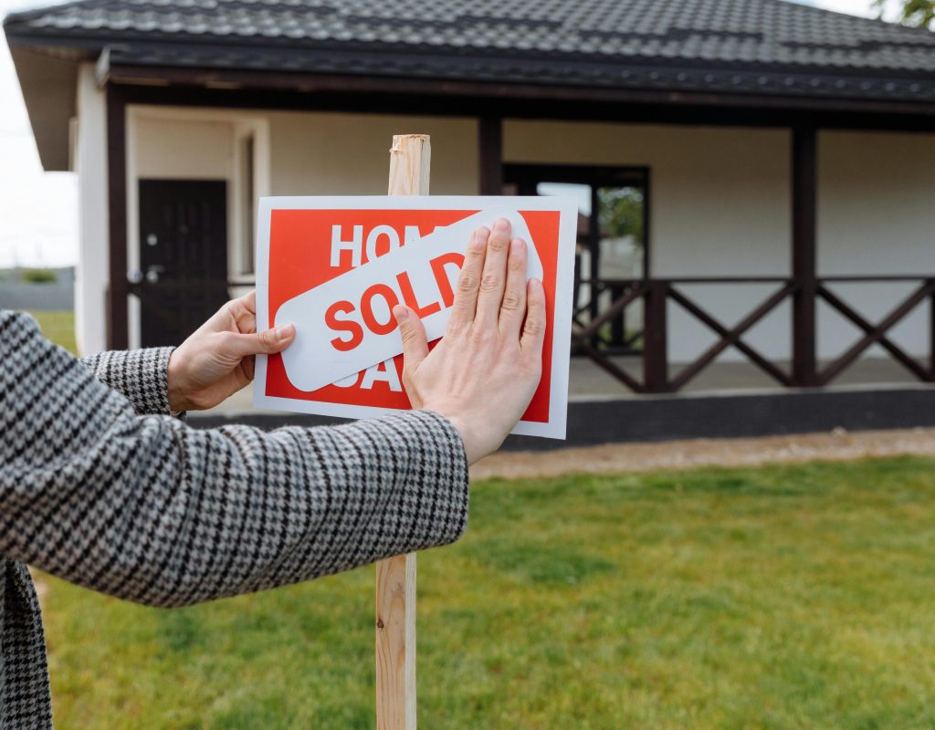 Close-up of hands placing 'sold' sticker on sign in front of a new house.